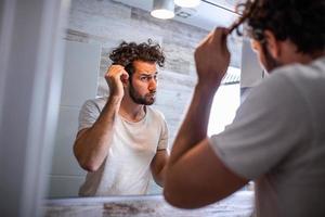 apuesto joven tocándose el pelo con la mano y acicalándose en el baño en casa. hombre metrosexual blanco preocupado por la pérdida de cabello y mirando al espejo su línea de cabello que retrocede. foto