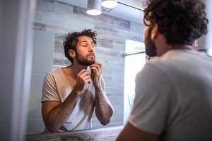 Reflection of handsome man with beard looking at mirror and touching face in bathroom grooming photo