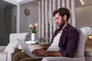 Man using laptop computer, working at home. Worried about finances. Handsome man with beard working at home on project, he is sitting on sofa looking at his laptop in front of him. Focus on the man photo