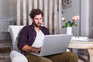 Man using laptop computer, working at home. Worried about finances. Handsome man with beard working at home on project, he is sitting on sofa looking at his laptop in front of him. Focus on the man photo