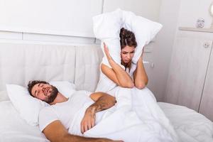 Snoring man. Couple in bed, man snoring and woman can not sleep. Picture of angry woman in bed with snoring man. Portrait of woman blocking ears with pillow while man snoring on bed photo