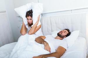 Young couple have problem with man's snoring. Heterosexual couple in bed, man sleeps and snoring with mouth open, while a tired woman irritated by snoring sitting on bed with a pillow on her head. photo