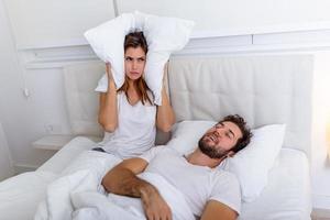 Young couple have problem with man's snoring. Heterosexual couple in bed, man sleeps and snoring with mouth open, while a tired woman irritated by snoring sitting on bed with a pillow on her head. photo