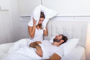 Young couple have problem with man's snoring. Heterosexual couple in bed, man sleeps and snoring with mouth open, while a tired woman irritated by snoring sitting on bed with a pillow on her head. photo
