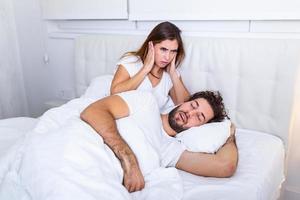 Man snoring while his wife is covering ears with hands. Woman suffers from her male partner snoring in bed. Noise concept. Real people. Young irritated woman lying in bed with snoring husband photo