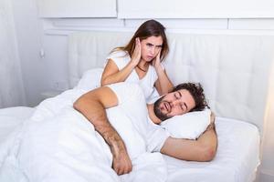 hombre roncando mientras su esposa cubre los oídos con las manos. la mujer sufre porque su pareja masculina ronca en la cama. concepto de ruido gente real. joven mujer irritada acostada en la cama con el marido roncando foto