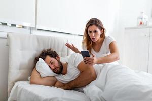 Jealous wife spying the phone of her partner while he is sleeping in a bed at home. Shocked jealous wife spying the phone of her husband while man sleeping in bed at home photo