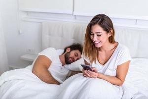 mujer mirando su teléfono mientras el hombre duerme en la cama. joven hermosa mujer hablando en privado por teléfono móvil mientras su marido duerme en la cama en el dormitorio