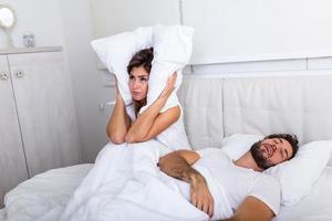 la pareja joven tiene problemas con los ronquidos del hombre. pareja heterosexual en la cama, el hombre duerme y ronca con la boca abierta, mientras que una mujer cansada irritada por los ronquidos sentada en la cama con una almohada en la cabeza. foto