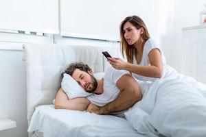 esposa celosa espiando el teléfono de su pareja mientras él duerme en una cama en casa. Sorprendida esposa celosa espiando el teléfono de su esposo mientras el hombre duerme en la cama en casa foto