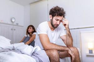 Beautiful girl and a frustrated man sitting in bed and not looking at each other. Upset couple ignoring each other. Worried man in tension at bed. Young couple angry with each other after a fight. photo