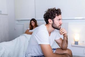 triste hombre pensativo después de discutir con novia. dificultades de relación, conflicto y concepto familiar, pareja infeliz que tiene problemas en el dormitorio. chico triste sentado en la cama, novia en el fondo foto