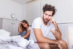 Young couple having a problem. Handsome man is leaning on his hand while sitting sadly on bed, woman is sitting in the background. Head full of thoughts when she is around photo