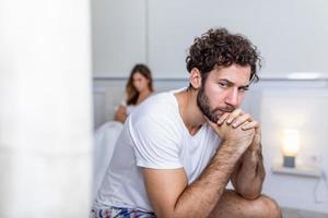 Sad thoughtful man after arguing with girlfriend.Relationship difficulties, conflict and family concept,unhappy couple having problems at bedroom. Sad guy sitting on bed,girlfriend in the background photo