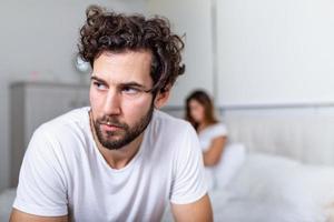 triste hombre pensativo después de discutir con novia. dificultades de relación, conflicto y concepto familiar, pareja infeliz que tiene problemas en el dormitorio. chico triste sentado en la cama, novia en el fondo foto