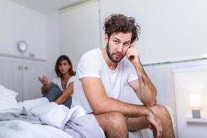 Young couple having a problem. Handsome man is leaning on his hand while sitting sadly on bed, woman is sitting in the background. Head full of thoughts when she is around photo