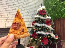 Hand held pizza slice with Christmas tree decoration background. photo