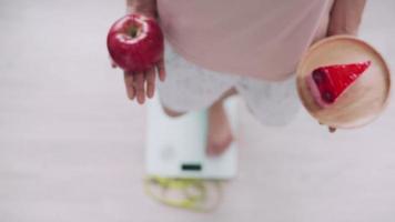 las mujeres están eligiendo los alimentos adecuados para una buena salud. las mujeres están ayunando. opciones de comparación entre pastel y manzanas durante la medición de peso en balanzas digitales. concepto de dieta video