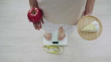 Women are choosing the right food for good health. Women are fasting. Comparison options between cake and  apples during weight measurement on digital scales. Diet concept video