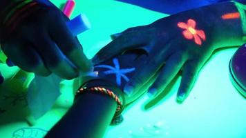 Nahaufnahme der Hand des kleinen Mädchens, die mit fluoreszierendem Malpulver bemalt wird. Auf dem Tisch liegen fluoreszierende Farbe und Bleistift. Body-Art-Konzept video