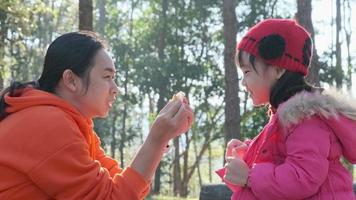 Happy mother and daughter eating bread for breakfast after camping in the pine forest. Portrait of happy mother and little daughter in winter park. Family winter vacation with children. video