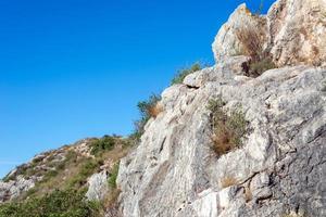 cracked rock covered with dry grass and plants on a backdrop of a hill and blue sky photo