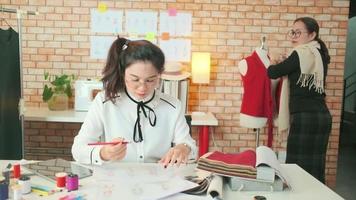una diseñadora de moda asiática de mediana edad le enseña a un joven sastre adolescente en estudio con hilo colorido y tela de costura para ideas de colección de diseño de vestidos, pequeña empresa boutique profesional. video