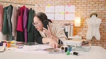 asiatico di mezza età femmina moda progettista lavori nel studio di parlando su mobile Telefono di idee e disegno schizzi per vestito design collezione ordini. professionale boutique sarto pmi imprenditore. video