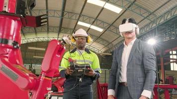 Afrikaanse Amerikaans ingenieur demonstreert robot arm controle naar klant met vr headsets simulator in mechanisch vervaardiging fabriek, intelligent industrieel machines, en futuristische technologie. video