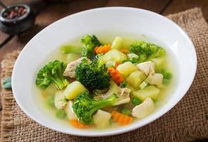 sopa de pollo con brócoli, guisantes, zanahorias y apio en un bol blanco sobre un fondo de madera de estilo rústico foto