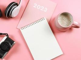 flat lay of blank paper note book on pink diary or planner 2023, headphones, camera  and pink cup of coffee on pink background. photo