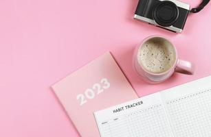 flat lay of habit tracker book on pink diary or planner 2023, pink cup of coffee and digital camera on pink background with copy space. photo