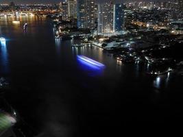 toma nocturna, larga exposición, luces iluminadas, ángulo alto, vista aérea del horizonte, paisaje urbano, orilla del río, río chao phraya, luna en el fondo, bangkok, tailandia foto