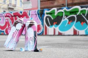 varias latas de aerosol usadas con pintura rosa y blanca y tapas para rociar pintura bajo presión se encuentran en el asfalto cerca de la pared pintada en dibujos de graffiti de colores foto