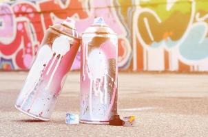 Several used spray cans with pink and white paint and caps for spraying paint under pressure is lies on the asphalt near the painted wall in colored graffiti drawings photo