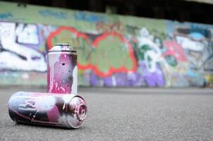 A few used spray cans with pink and white paint lie on the asphalt against the background of a painted wall in colorful graffiti drawings photo