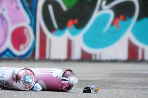 Several used spray cans with pink and white paint and caps for spraying paint under pressure is lies on the asphalt near the painted wall in colored graffiti drawings photo