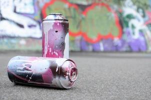 A few used spray cans with pink and white paint lie on the asphalt against the background of a painted wall in colorful graffiti drawings photo