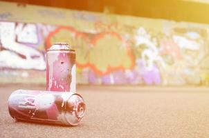 A few used spray cans with pink and white paint lie on the asphalt against the background of a painted wall in colorful graffiti drawings photo