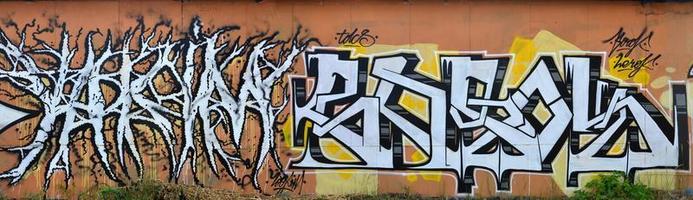 A photo of several graffiti artworks on the metal wall. Graffiti drawings are made with white paint with black outlines and have an orange background. Texture of wall with graffiti decoration