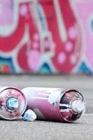 Several used spray cans with pink and white paint and caps for spraying paint under pressure is lies on the asphalt near the painted wall in colored graffiti drawings photo