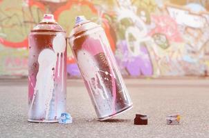 varias latas de aerosol usadas con pintura rosa y blanca y tapas para rociar pintura bajo presión se encuentran en el asfalto cerca de la pared pintada en dibujos de graffiti de colores foto