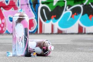 Several used spray cans with pink and white paint and caps for spraying paint under pressure is lies on the asphalt near the painted wall in colored graffiti drawings photo