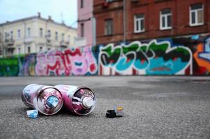 Several used spray cans with pink and white paint and caps for spraying paint under pressure is lies on the asphalt near the painted wall in colored graffiti drawings photo