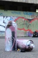 Several used spray cans with pink and white paint and caps for spraying paint under pressure is lies on the asphalt near the painted wall in colored graffiti drawings photo