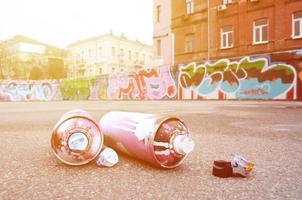 Several used spray cans with pink and white paint and caps for spraying paint under pressure is lies on the asphalt near the painted wall in colored graffiti drawings photo