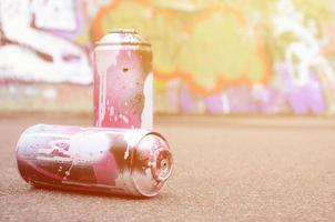 A few used spray cans with pink and white paint lie on the asphalt against the background of a painted wall in colorful graffiti drawings photo