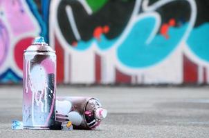 Several used spray cans with pink and white paint and caps for spraying paint under pressure is lies on the asphalt near the painted wall in colored graffiti drawings photo