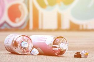 Several used spray cans with pink and white paint and caps for spraying paint under pressure is lies on the asphalt near the painted wall in colored graffiti drawings photo