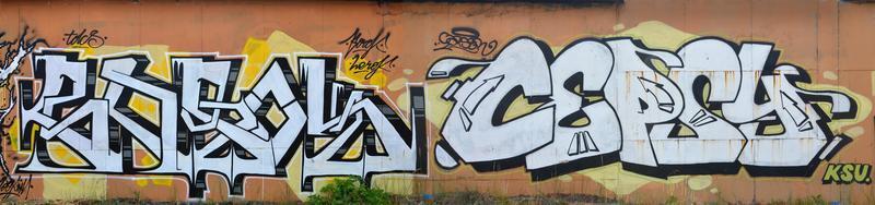 A photo of several graffiti artworks on the metal wall. Graffiti drawings are made with white paint with black outlines and have an orange background. Texture of wall with graffiti decoration